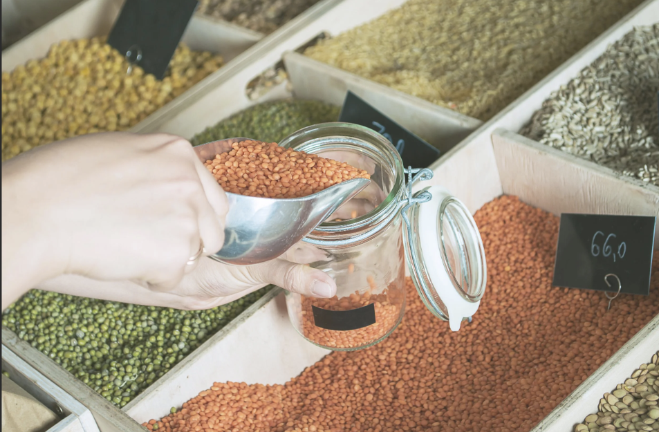 a hand with a metal scoop refills beans from a bin into a glass reusable jar